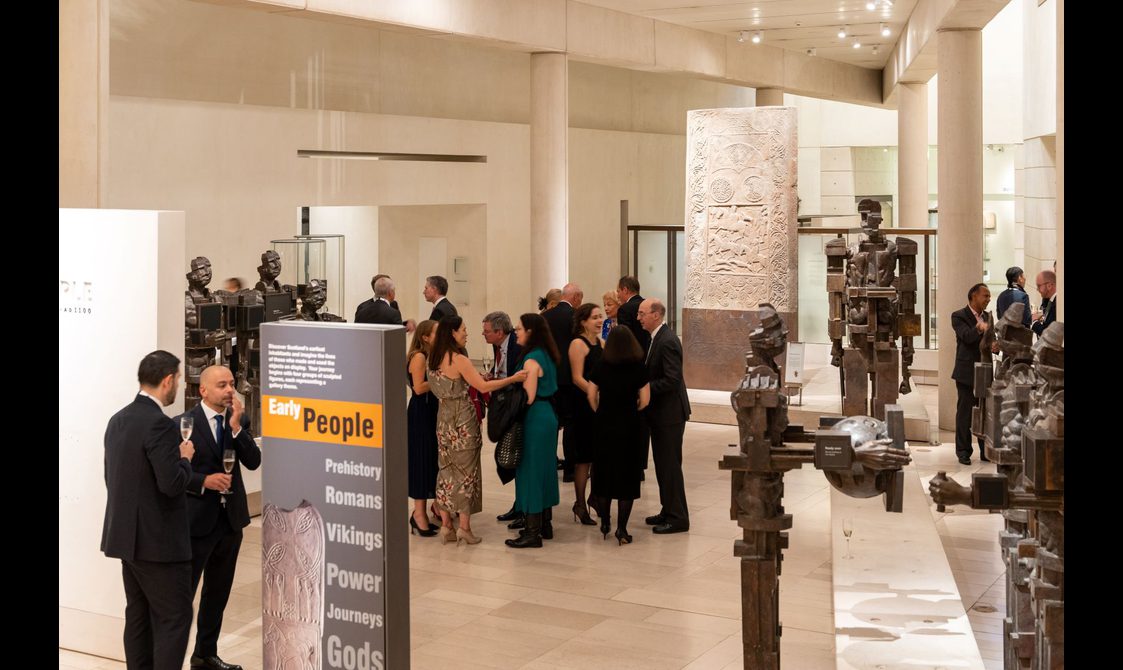 A group of people at a drinks reception in a museum gallery with white walls and high ceilings. Around the gallery are scultpures and artefacts from Scottish history.