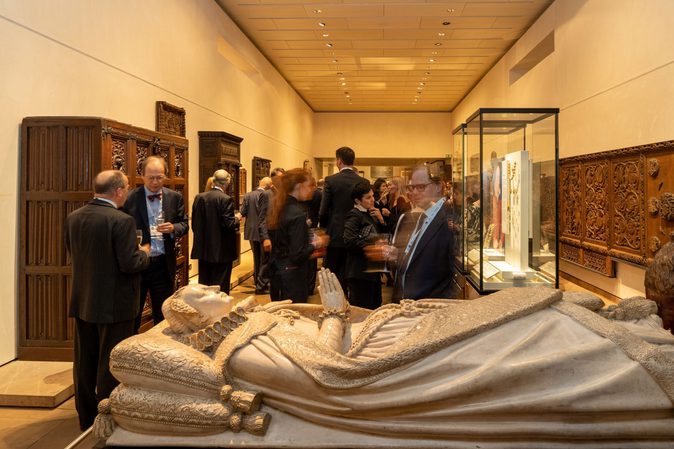 A group of people at a drinks reception in a museum gallery. At the front is a replica of the casket of Mary Queen of Scots. Lining the walls are ornate pieces of wooden furniture.