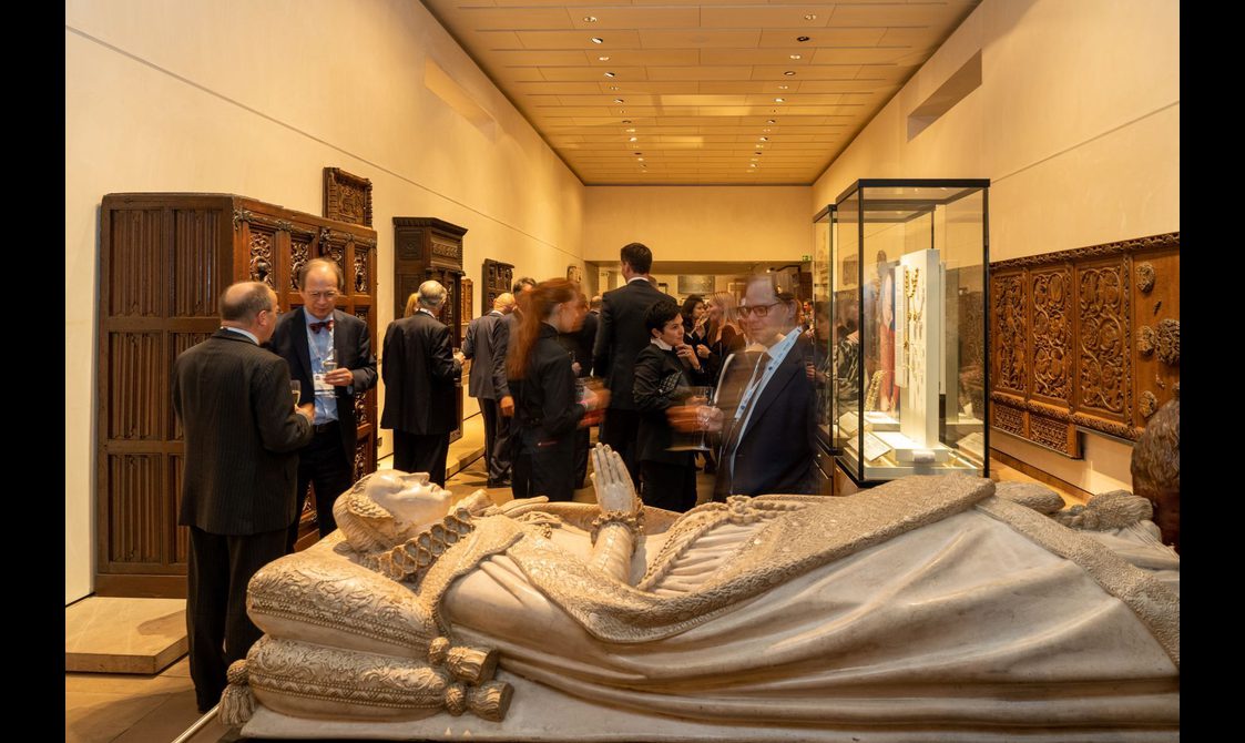 A group of people at a drinks reception in a museum gallery. At the front is a replica of the casket of Mary Queen of Scots. Lining the walls are ornate pieces of wooden furniture.