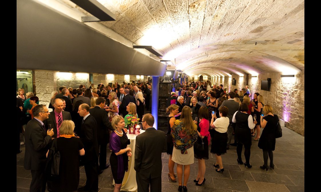 People at a drinks reception in a long hall with stone floors and a low arched ceiling.