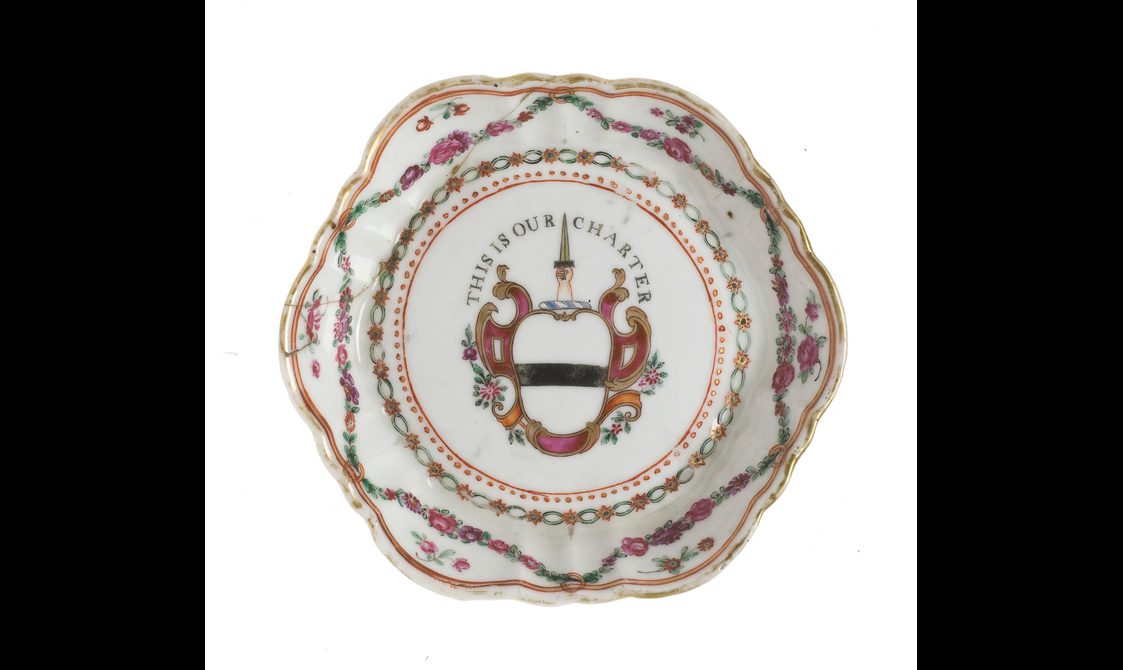 Hexagonal plate with red flower motifs around the edges of the plate, A red and black coat of arms with "This is our charter" phrase written in a crescent shape frames a hand holding a dagger reaching upwards in the center.