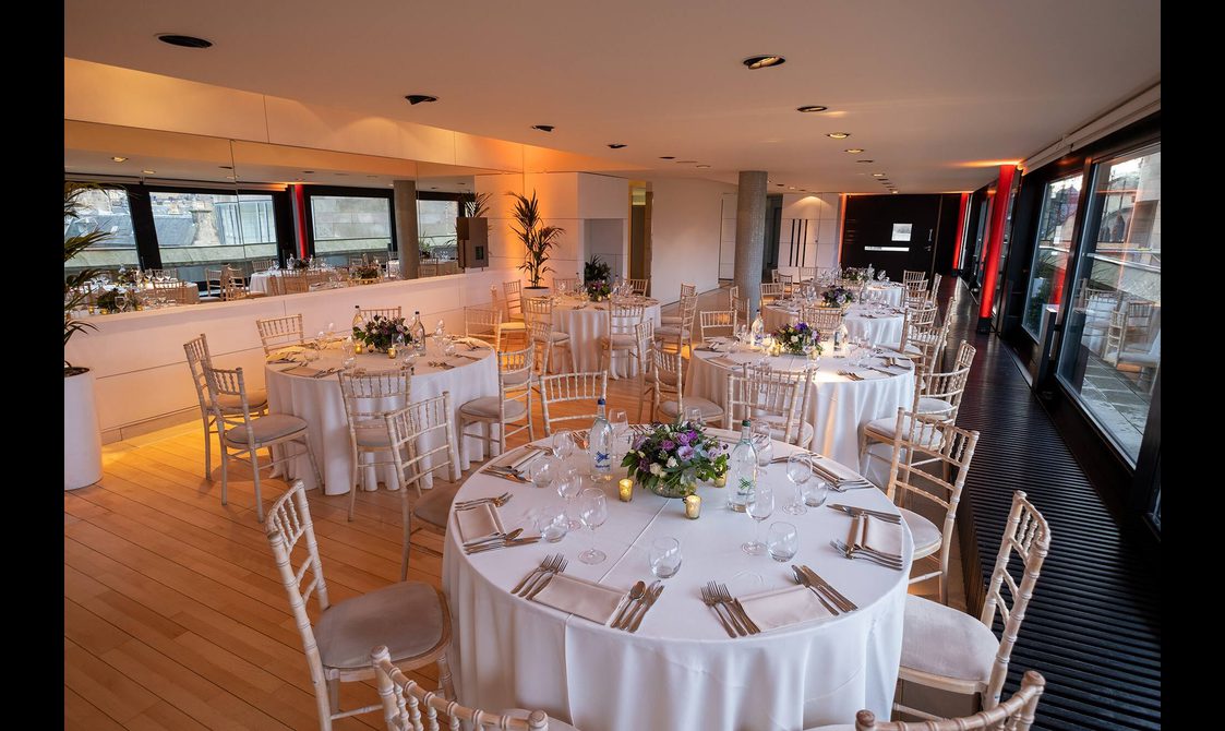 An event space with tables set up for a meal. The tables are covered with white sheets and there are rooftop views through the windows.