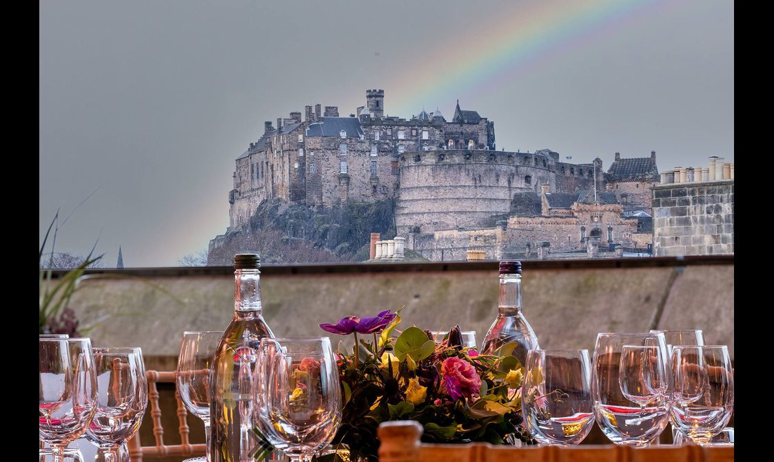 View of Edinburgh Castle from a rooftop space. A table is set out with wine glasses and decorated with flowers. There is a rainbow in the background..