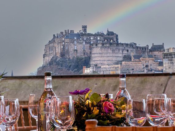 View of Edinburgh Castle from a rooftop space. A table is set out with wine glasses and decorated with flowers. There is a rainbow in the background..