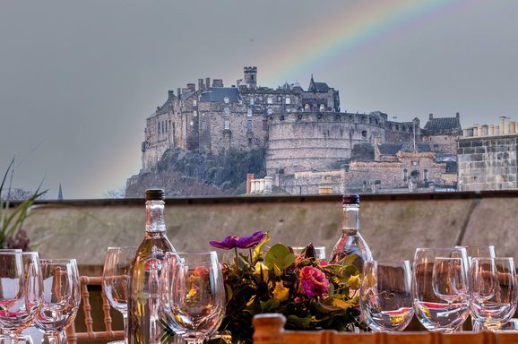 View of Edinburgh Castle from a rooftop space. A table is set out with wine glasses and decorated with flowers. There is a rainbow in the background..