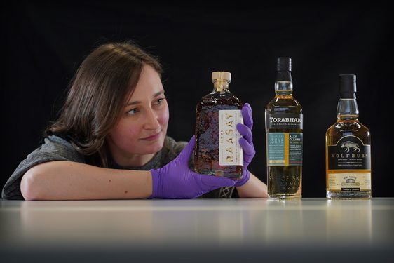 Laura Scobie leans on a white surface. She is looking at a whisky bottle she holds with purple latex gloved hands. There are two other bottles on the table.