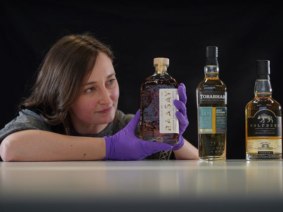 Laura Scobie leans on a white surface. She is looking at a whisky bottle she holds with purple latex gloved hands. There are two other bottles on the table.