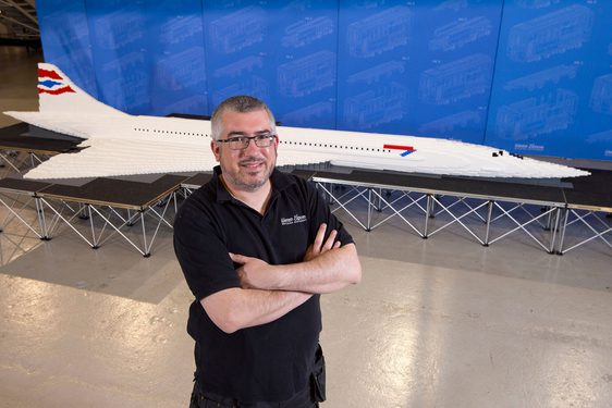 A person with there arms folded standing in front of a lego model of a concorde plane.