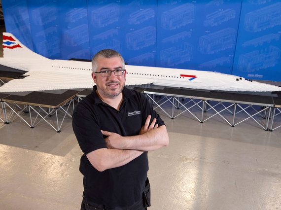A person with there arms folded standing in front of a lego model of a concorde plane.