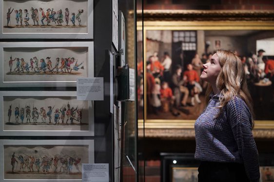 A person with long blonde hair standing in front of a museum case, with a large portrait in the background.
