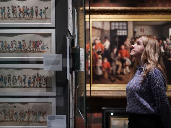 A person with long blonde hair standing in front of a museum case, with a large portrait in the background.