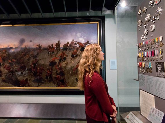 A visitor looking at a series of military medals on display in a museum case. There is a large portrait in the background depicting a battle.