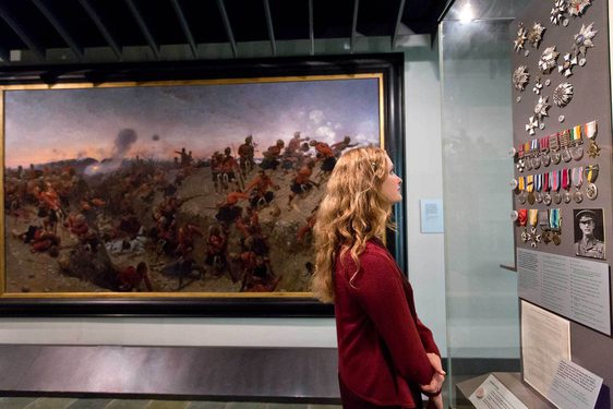 A visitor looking at a series of military medals on display in a museum case. There is a large portrait in the background depicting a battle.
