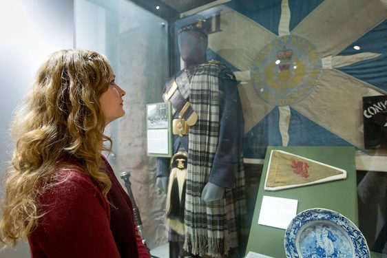 A visitor looking at a museum display case showcasing items related to Scottish military history. A mannequin in traditional Scottish dress stands in the case in front of a saltire flag.