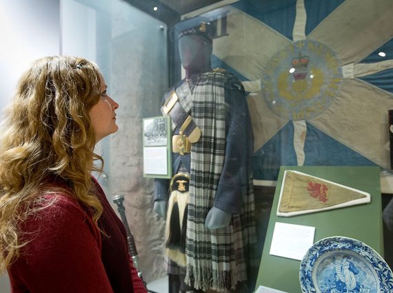 A visitor looking at a museum display case showcasing items related to Scottish military history. A mannequin in traditional Scottish dress stands in the case in front of a saltire flag.