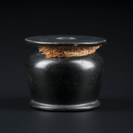 A black vase with a wide rim and a small opening. There is a piece of woven light brown fabric around its short neck.