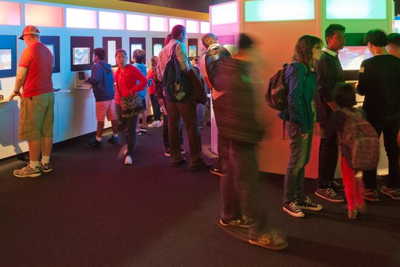 Ten players crowd around a line of consoles in an exhibition space at Game On in Ontario Science Centre.ario Science Ce