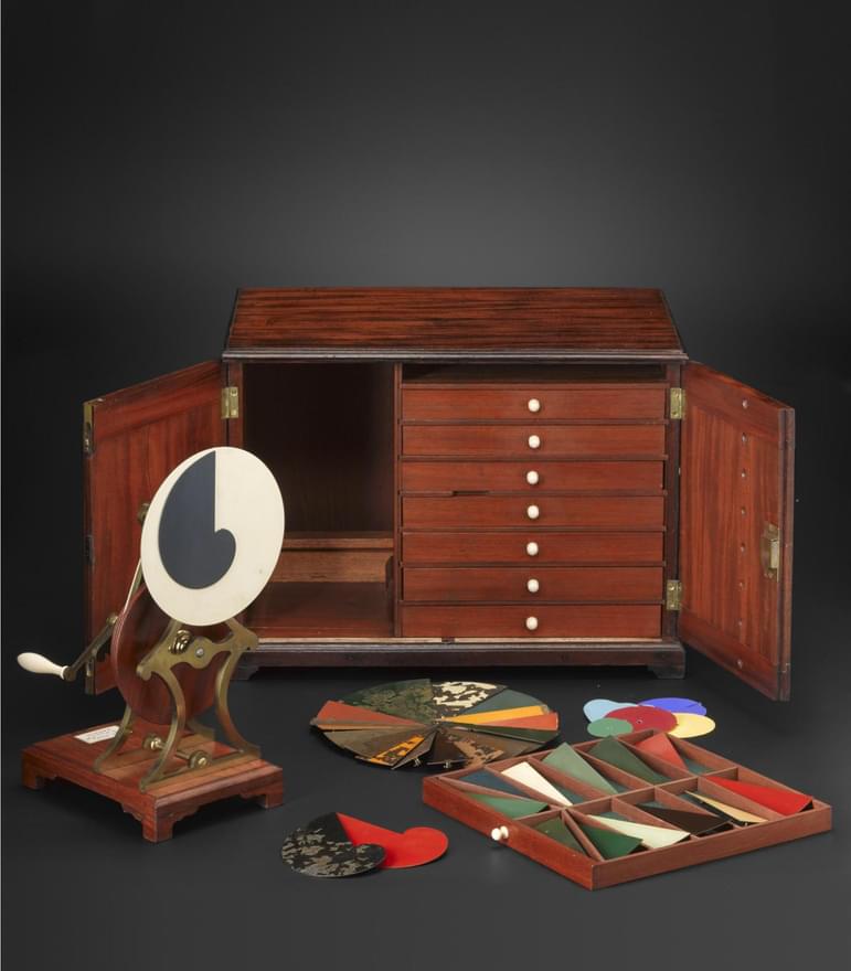 A wooden box with several small drawers with round pieces of coloured paper and a wheel on a small wooden stand in front of it.