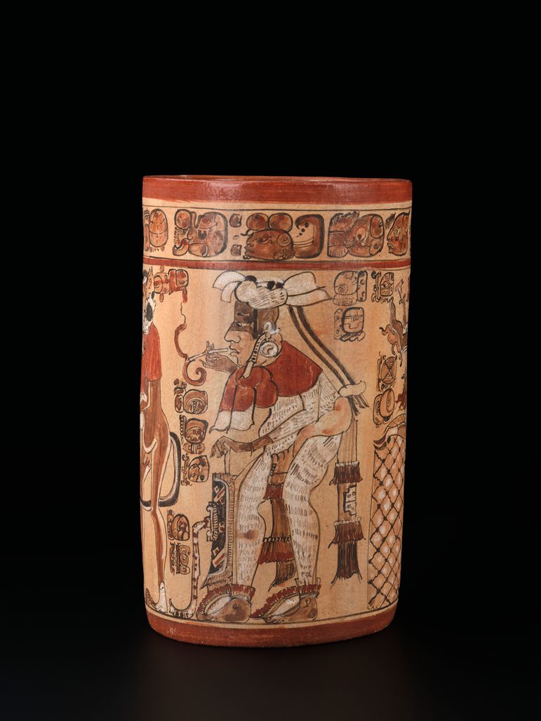 Ancient Mayan cylindrical vase displaying a high-status figure adorned with intricate regalia, surrounded by detailed hieroglyphics on a black background.
