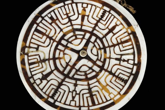 A circular shell pendant from Oceania with a white outer disk and brown inner disc. The brown disc is carved with intricate designs.