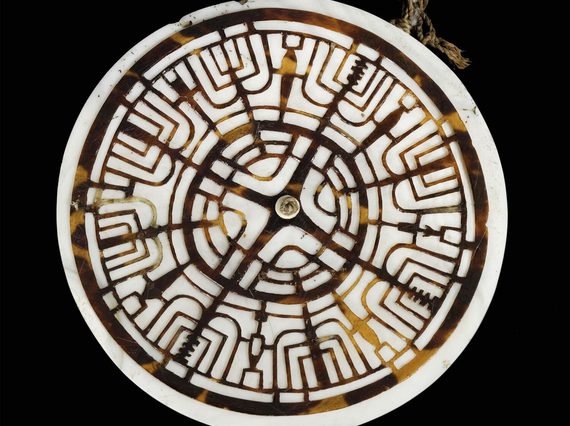 A circular shell pendant from Oceania with a white outer disk and brown inner disc. The brown disc is carved with intricate designs.
