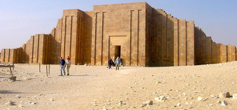 A large palace made of sandy coloured bricks with very angular details. The palace is in a desert and people can be seen in front of the palace.