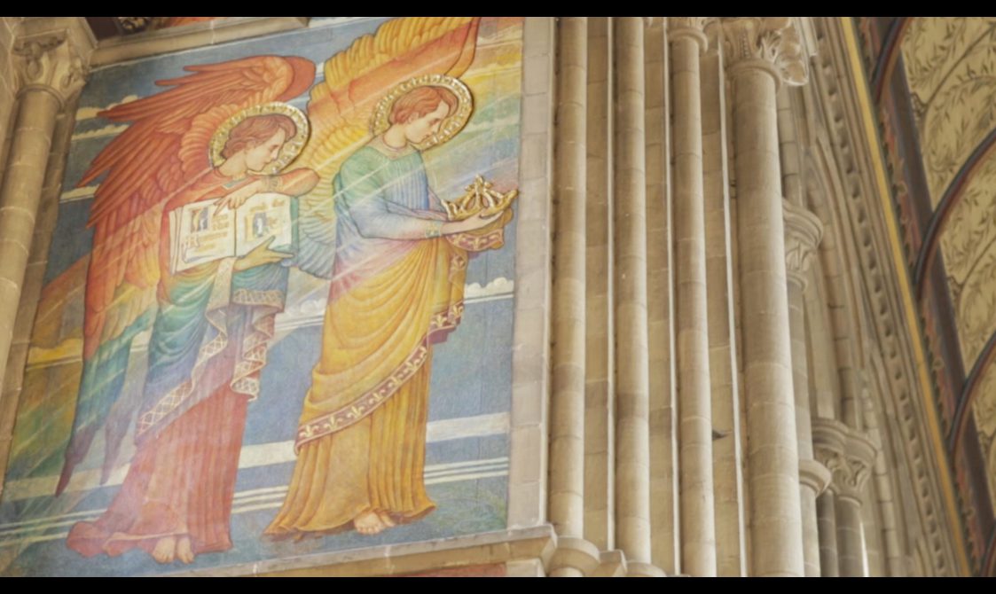 A mural in a church of two colourful angels, one is holding a book, the other is holding a crown.