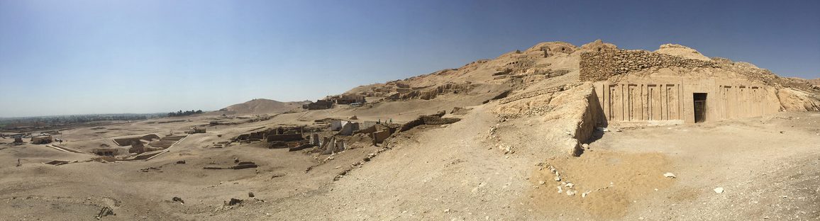 A panoramic view of a desert set against blue sky with a sand stone coloured building emerging to the right.