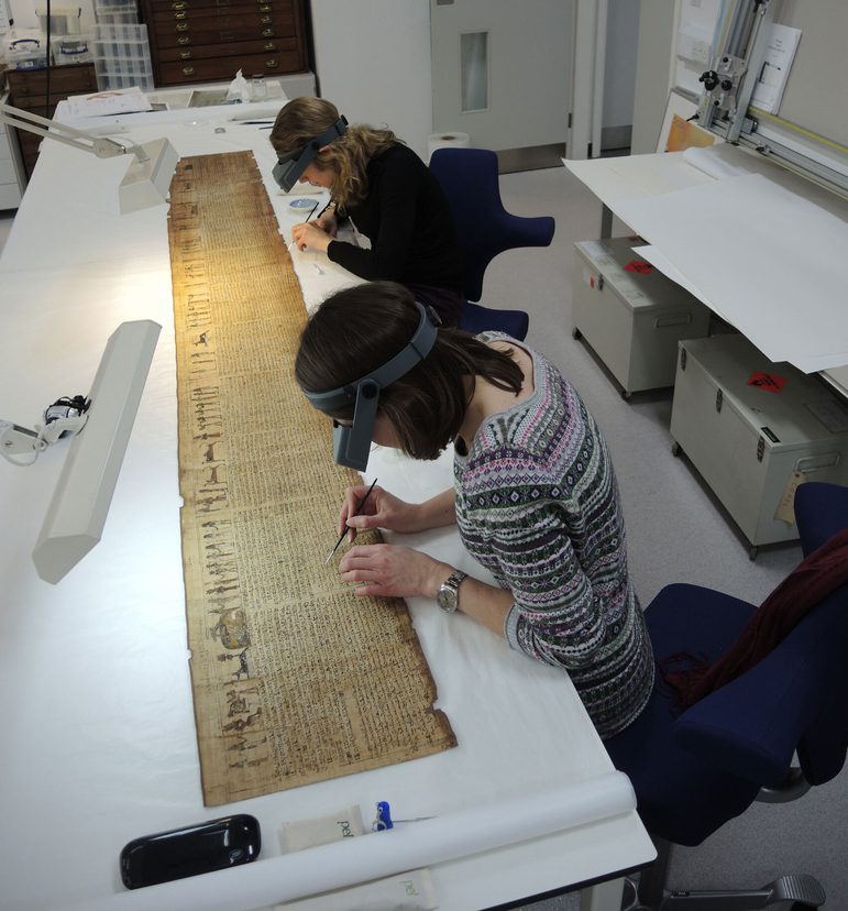 Two women sitting in office chairs wearing headgear with magnifying glasses and lights sit at a long write table working on a long ancient Egyptian scroll.