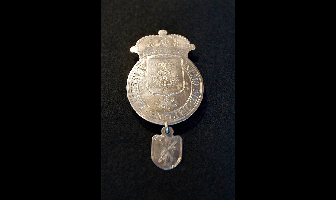 A round-shaped silver badge with engraved letters to the edges, a shield shaped Scottish thistle decoration in the center. An engraved crown attached to the top. A shield-shaped medallion with St Andrew decoration attached to the bottom.