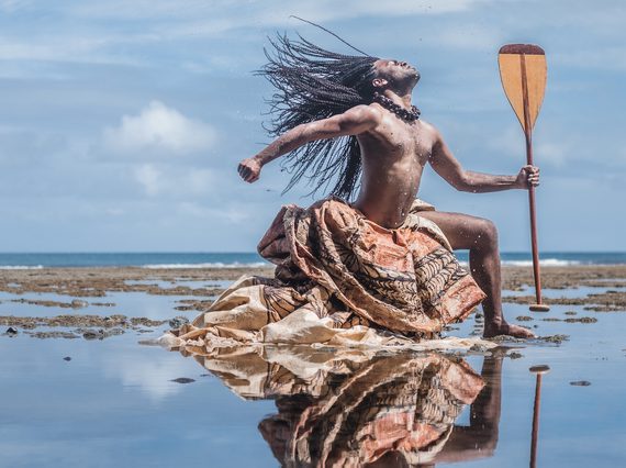 A man from Oceania dramatically poses on a beach covered in a shallow layer of water and seaweed. Throwing his head back and striking a 'warrior pose', his long braided hair, traditional dyed cloth wrapping below his waist, and the boat paddle he holds make him seem powerful and heroic.