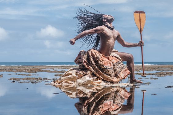 A man from Oceania dramatically poses on a beach covered in a shallow layer of water and seaweed. Throwing his head back and striking a 'warrior pose', his long braided hair, traditional dyed cloth wrapping below his waist, and the boat paddle he holds make him seem powerful and heroic.