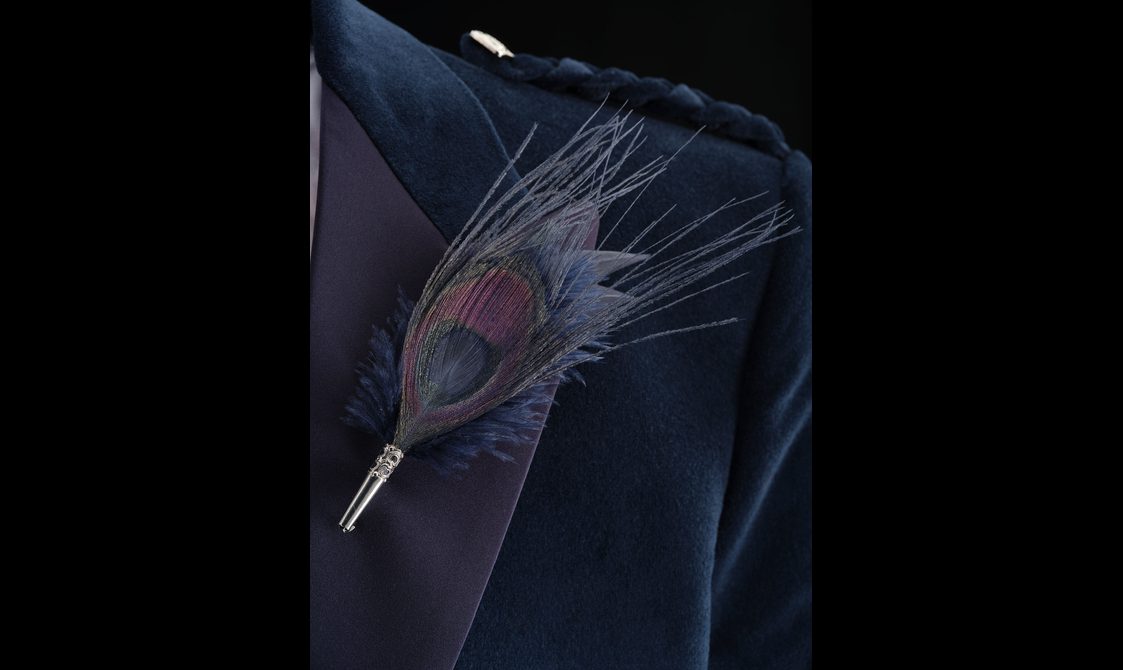 A detail of a navy blue velvet jacket with a peacock feather pin.