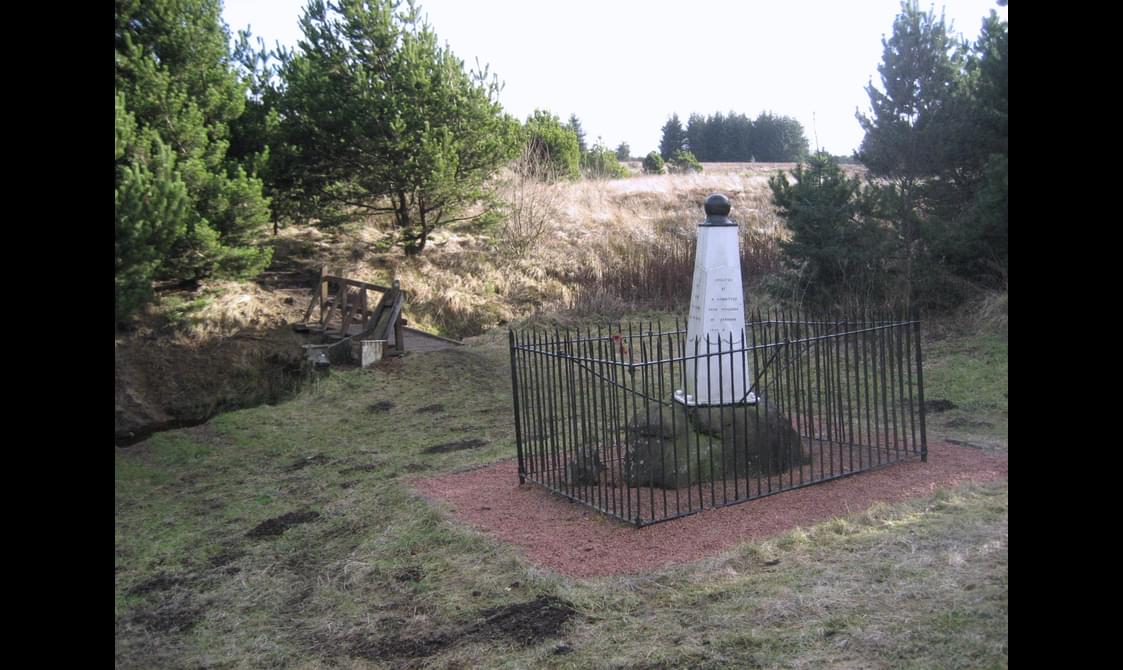A monument made up of a white column sitting on a rock with a fence around it in and countryside.