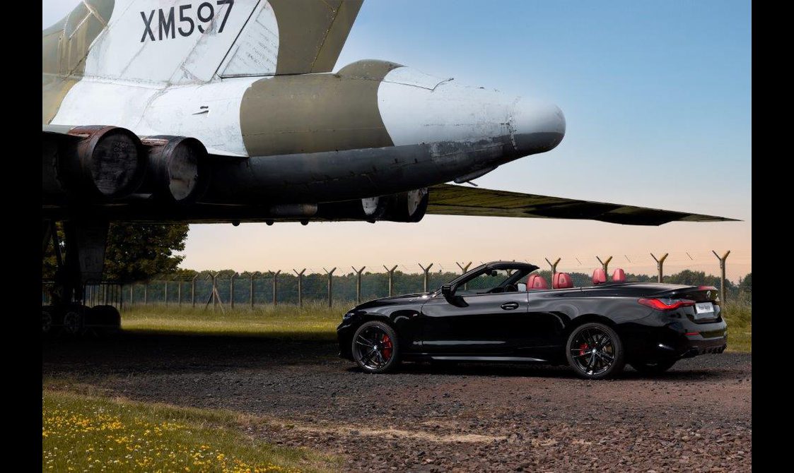 A convertible car with the top down parked outside under the tail of a military airplane.