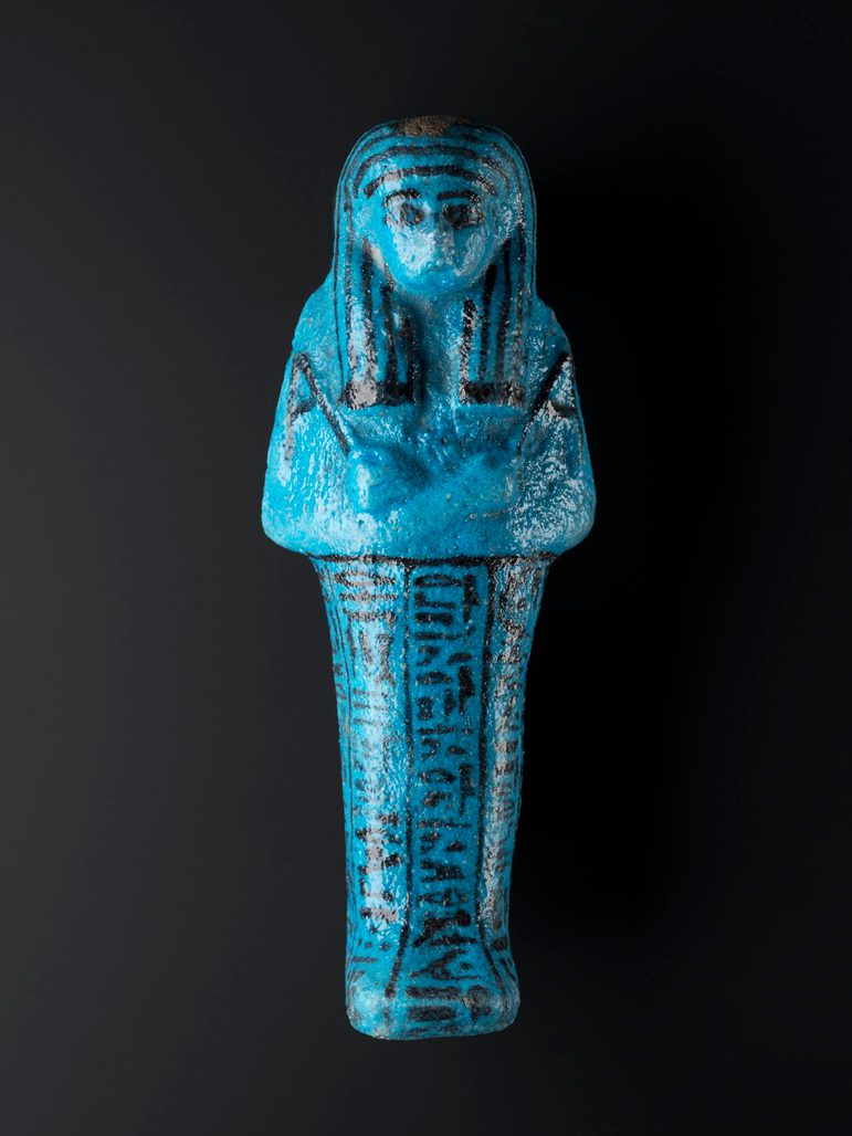 A ceramic blue figure made in the shape of a mummified person with detailed features and inscriptions. The figure is against a black background.