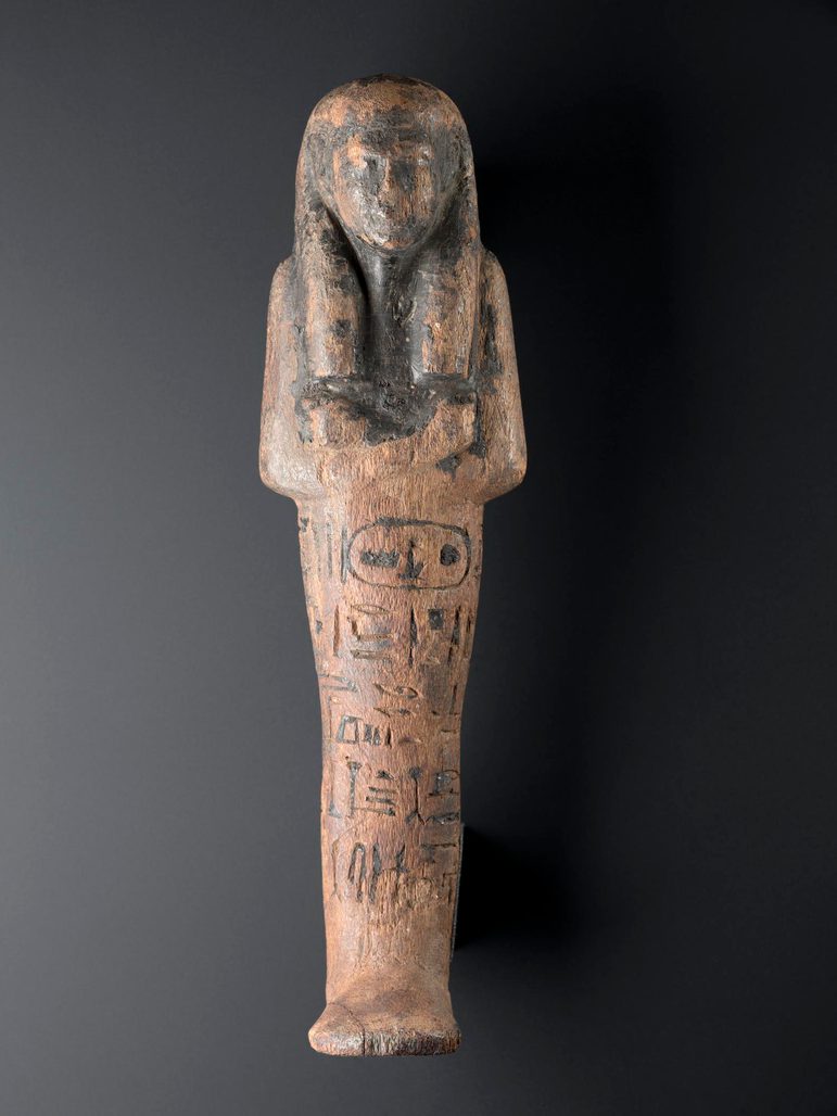 A light wooden figure in the shape of a mummified person with inscription and traces of black resin. The figure is against a black background.