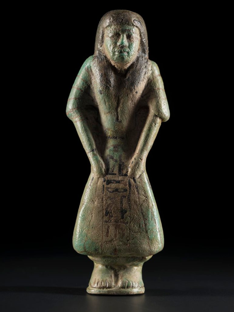 A dark green ceramic figure in a contemporary clothing of Egypt in the 1295-1069 BC. The figure is in the shape of a mummified person with inscriptions, and is against a black background.
