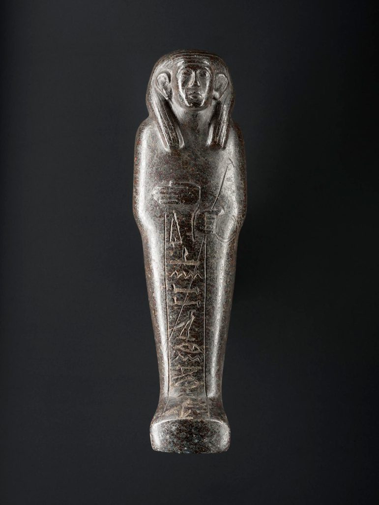 A black stone figure in the shape of a mummified person, with a large hieroglyphic ankh symbol and a sceptre. The figure is against a black background.