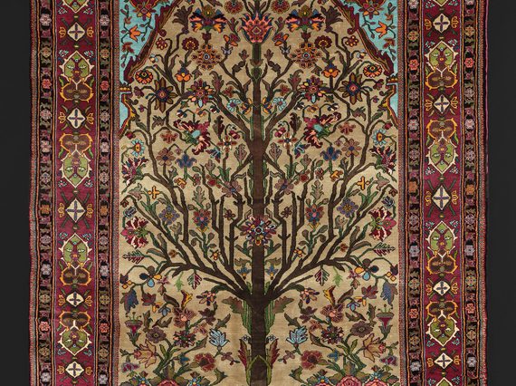 Silk rug depicting an angular tree with branches full of flowers. It is surrounded by a red border with various embellishments.