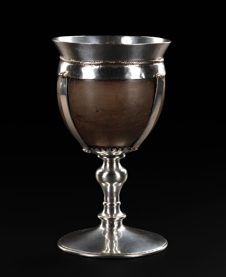 A colour photograph of a coconut cup on. black background. It has a tall decorative silver stem with a silver rim around the top and silver braces connecting the rim and the stem.