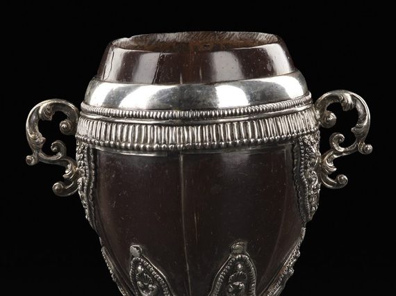 A colour photograph of an ornate cup made from a polished coconut shell with very elaborate silver embellishment of a rim, handles and stem. The cup has a coconut lid. Above the base is a set if silver designs of Serpens or dragons.