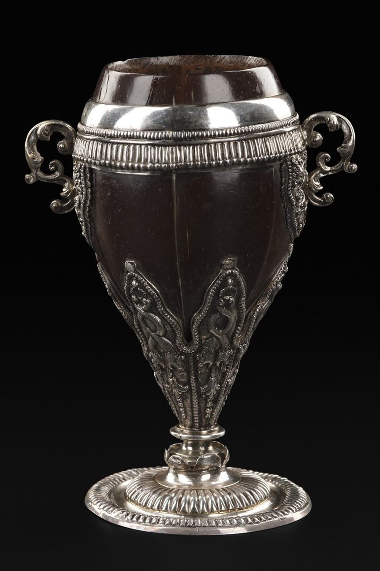 A colour photograph of an ornate cup made from a polished coconut shell with very elaborate silver embellishment of a rim, handles and stem. The cup has a coconut lid. Above the base is a set if silver designs of Serpens or dragons.