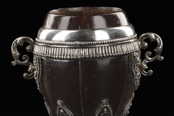 A colour photograph of an ornate cup made from a polished coconut shell with very elaborate silver embellishment of a rim, handles and stem. The cup has a coconut lid. Above the base is a set if silver designs of Serpens or dragons.