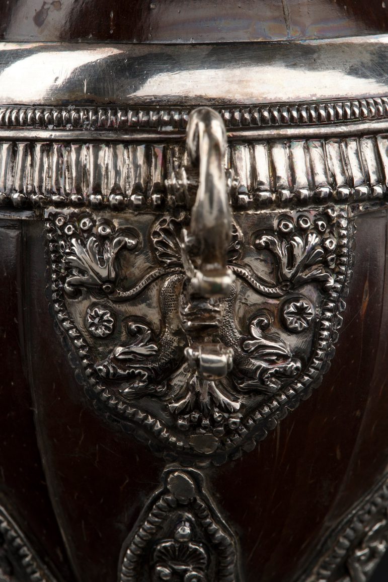 A colour photograph of a close up of the handle area of a silver embossed coconut cup. There is a vertical pattern across the whole rim where the hand meets the cup. Underneath the handle are snakes or dragon heads with elongated tails that have leaf shapes at the end.