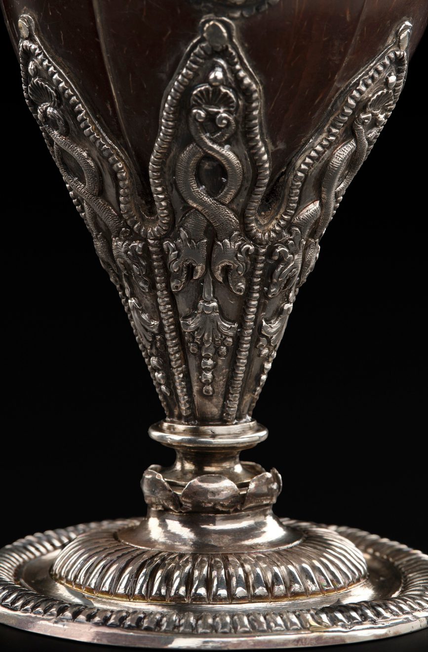 A colour photograph of a close up of the base of an ornate cup made from a polished coconut shell with very elaborate silver embellishment on the stem. Above the base is a set of silver designs of serpents or dragons.