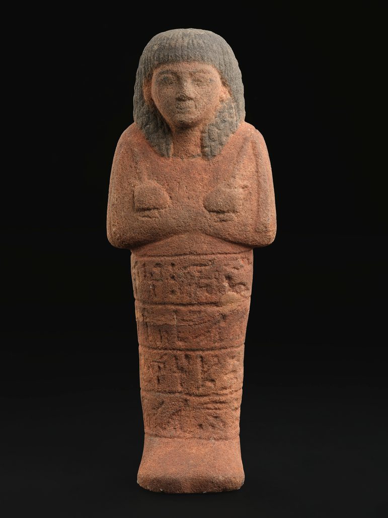 A large terracotta orange red sandstone figure in the shape of a mummified person with inscriptions. The figure is against a black background.