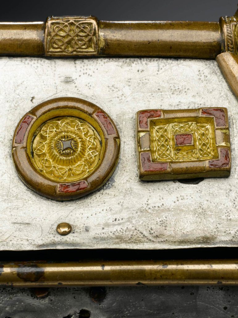 Close up of the roof of a silver box with gold metalwork. In focus are mounts of different shapes, each with interlaced designs.