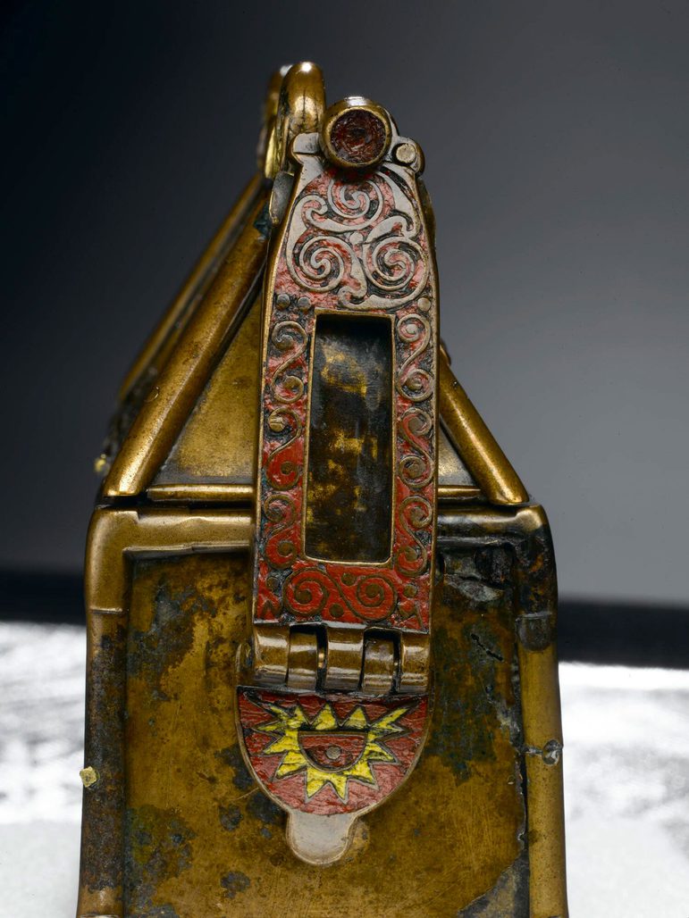 Side view of a small elaborate metal box. In focus is a decorated side plate painted a deep red and engraved with swirling patterns.