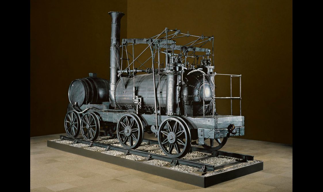A large black locomotive from the 19th century on display against a brown background.
